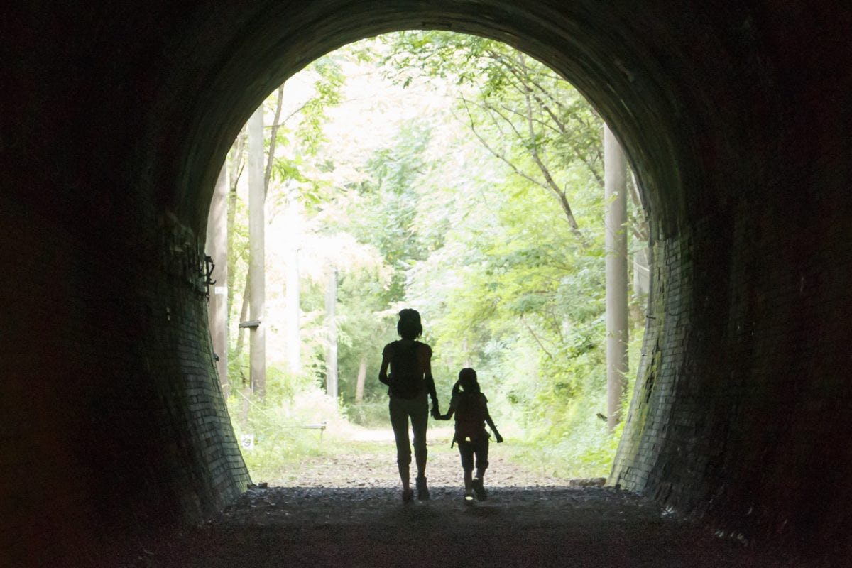 A tunnel looking out into a forested area. Two figures, one adult, one child, are silhouetted and holding hands 