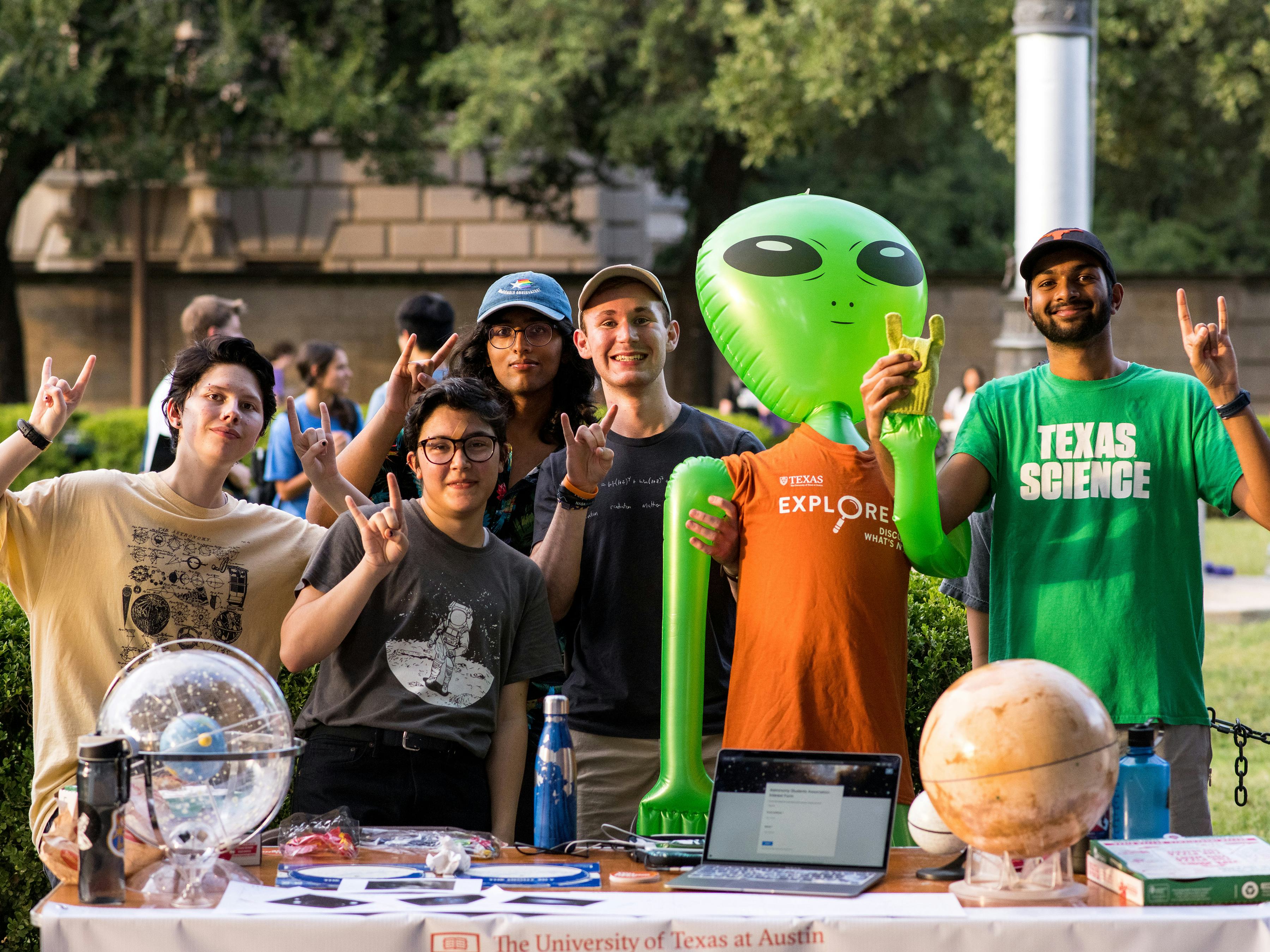 Physical sciences students tabling on the main mall. The students are gesturing Hook 'em hand gestures and posing with an inflatible green alien.