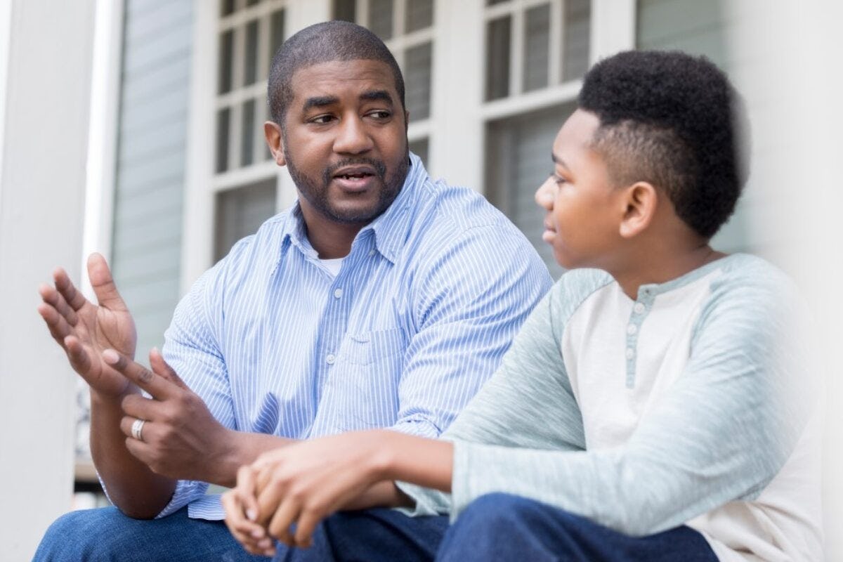 A Black man, left sits beside a Black boy on a front porch as the two talk