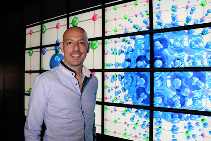 Giustino stands in front of a wall of LCD screens displaying computational models