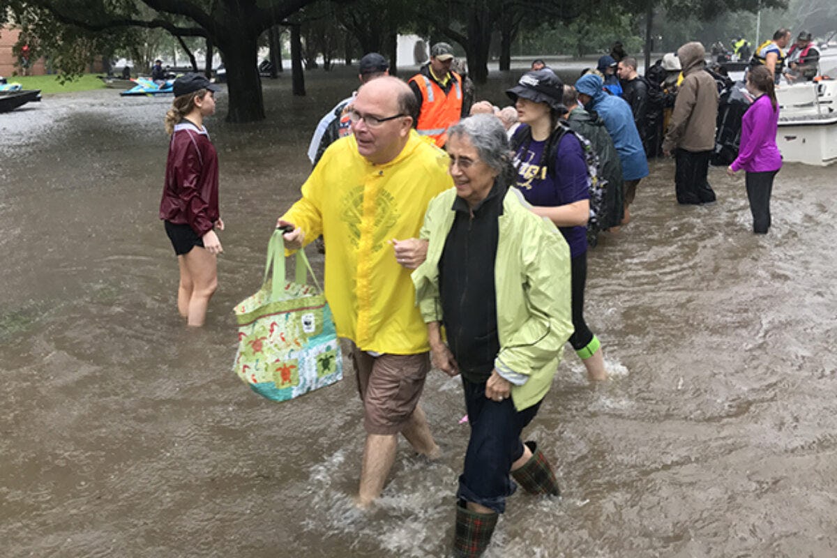 An older man in a yellow rain coat holds the arm of an older woman, right, as they wade through ankle-deep flood waters during Hurricane Harvey