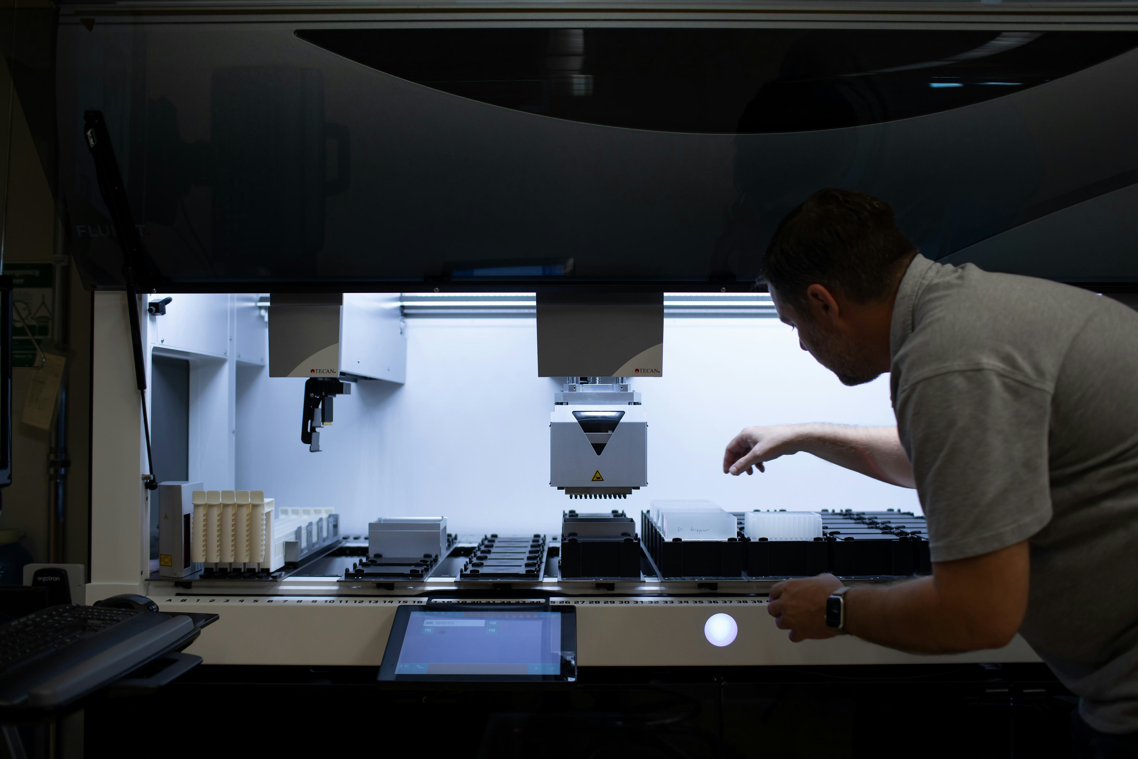 A scientist leans into a laboratory hood and moves samples under a robotic arm