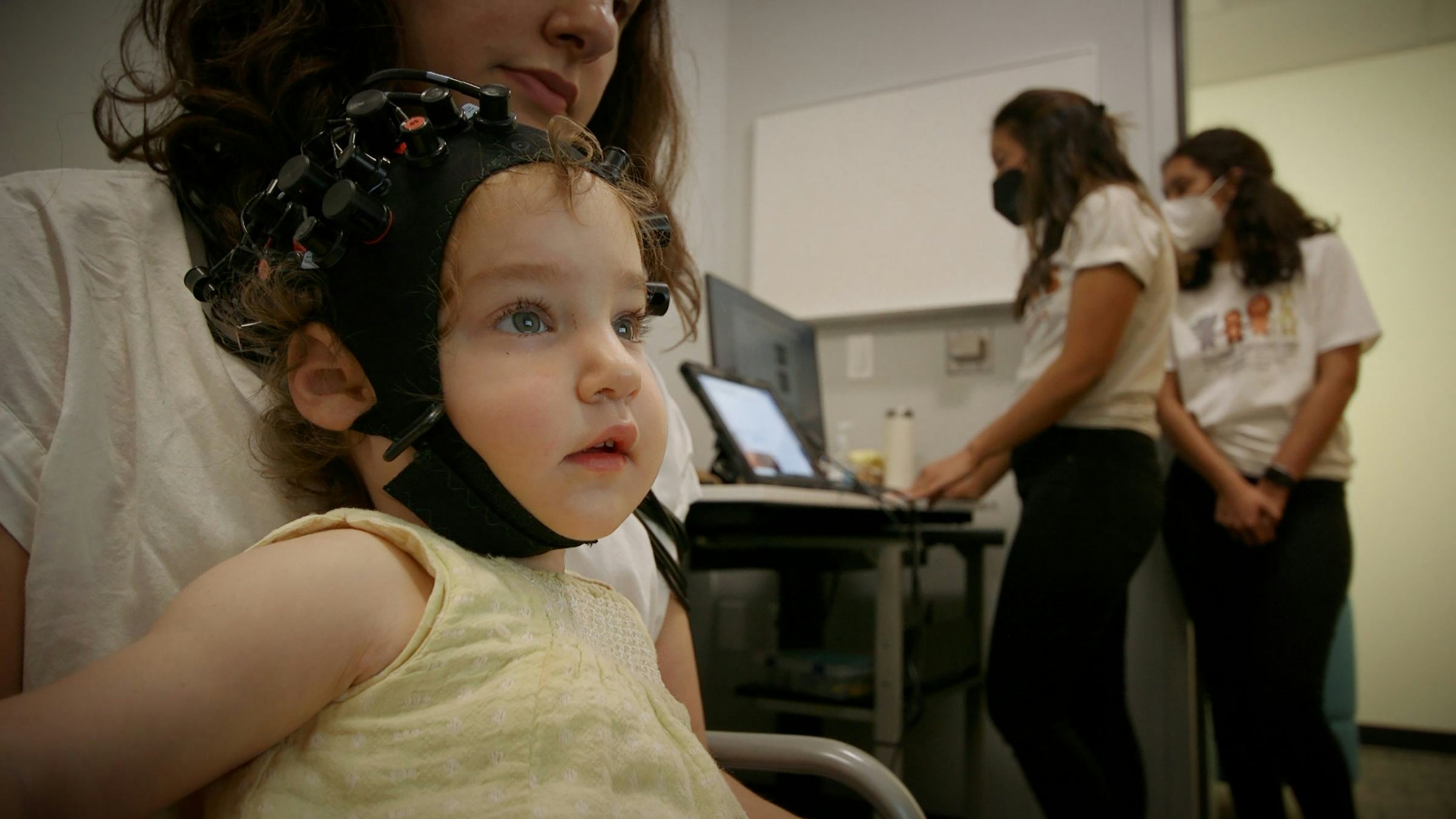 Girl wearing a black cap and sitting on her mother's lap looks off camera while two scientists look at a computer in the background