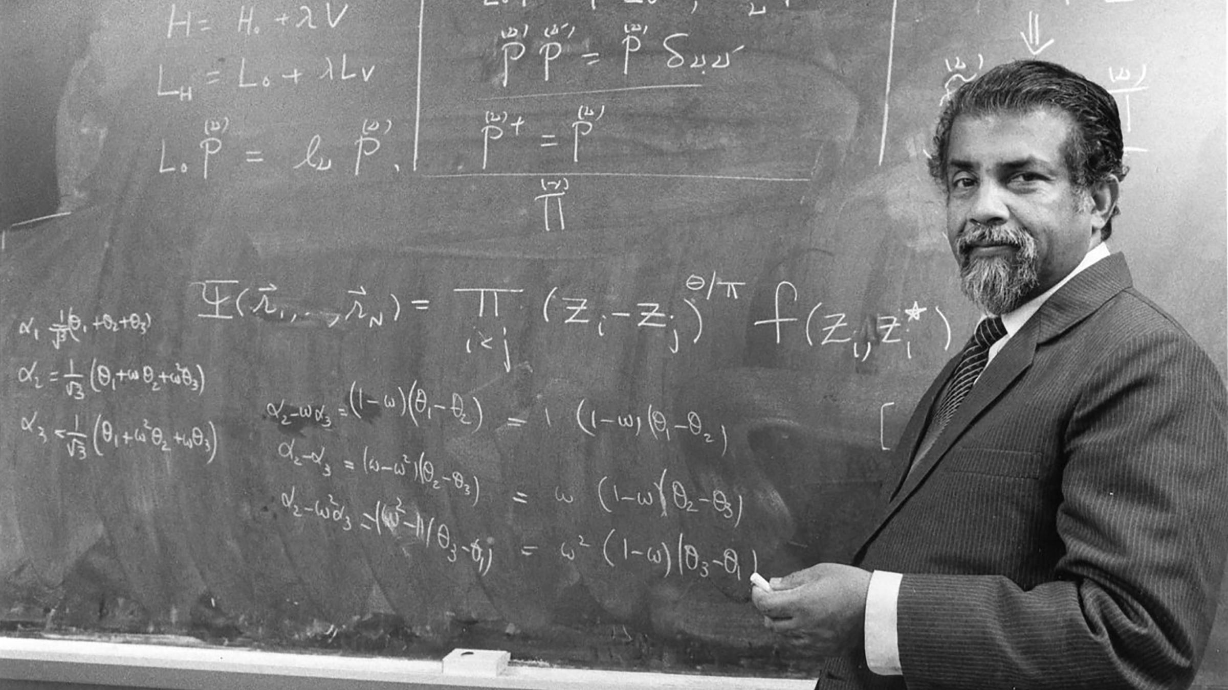 A man in a suit stands at a chalkboard bearing mathematical notation