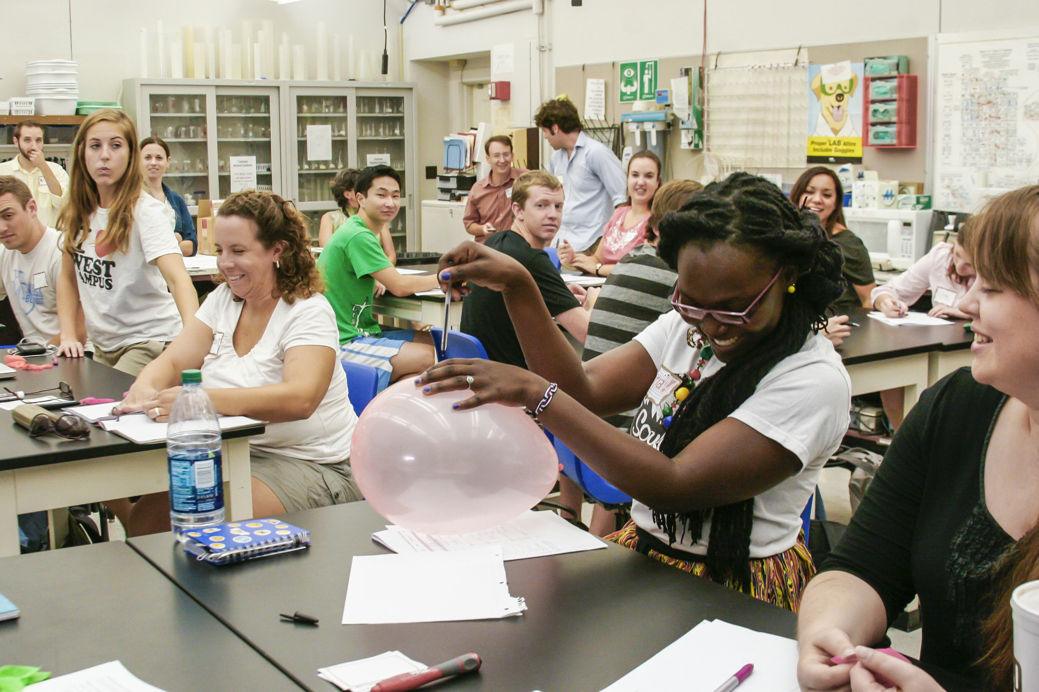 A young woman in a class practices an experiment with a balloon and looks afraid it will pop while peers look on