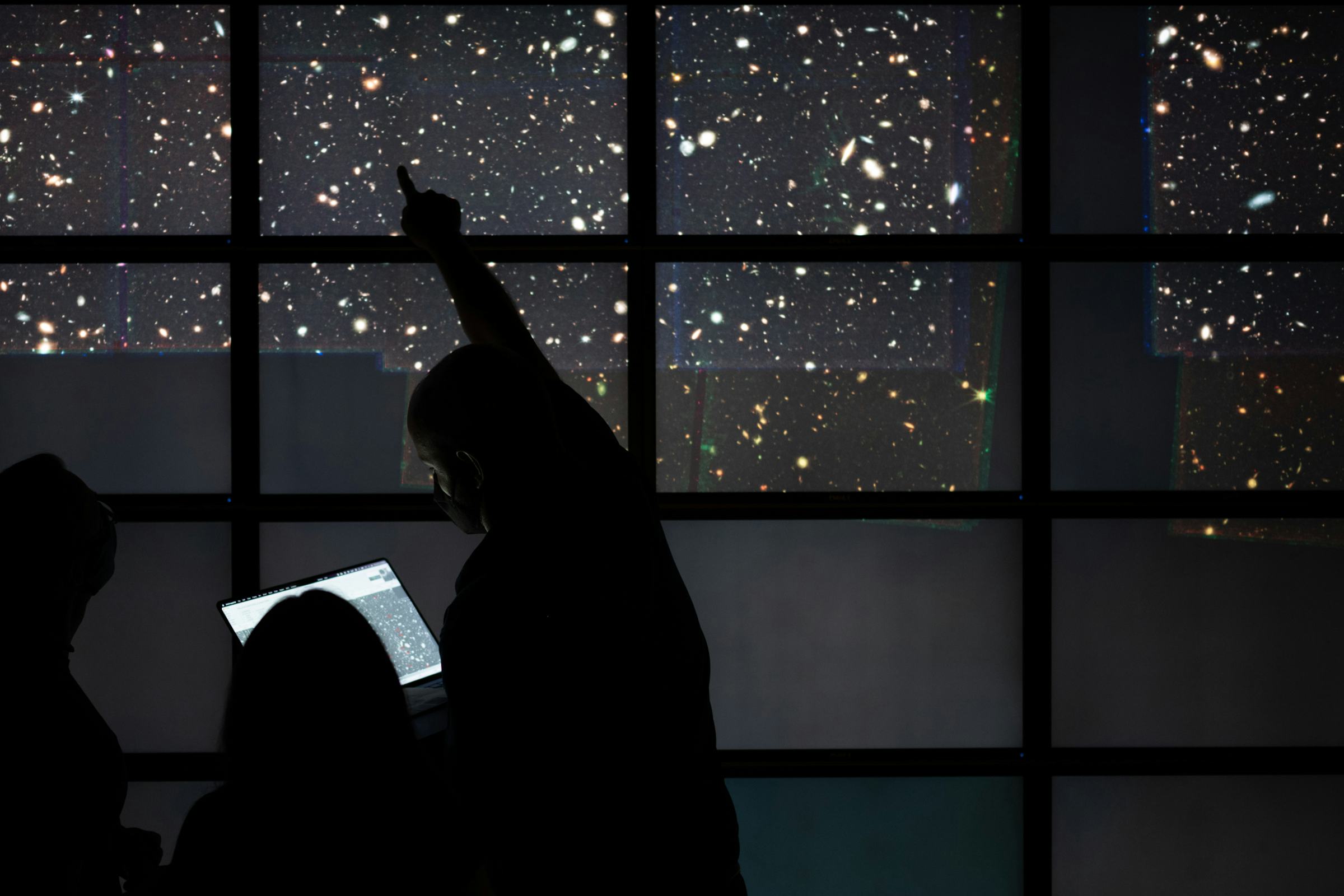 Silhouettes of three people in front of a large display of stars and galaxies, one person pointing at a specific spot